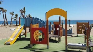 Une aire de jeux avec un toboggan, des structures d’escalade et une balançoire, le tout situé sur une plage.