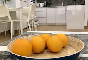 Una mesa de comedor con un cuenco de naranjas y una cafetera al fondo.