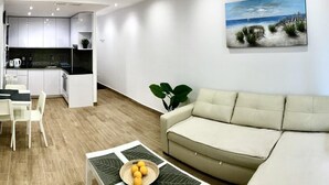 A modern living room with a sofa, a dining table with chairs, a kitchen area, and a beach-themed painting on the wall.