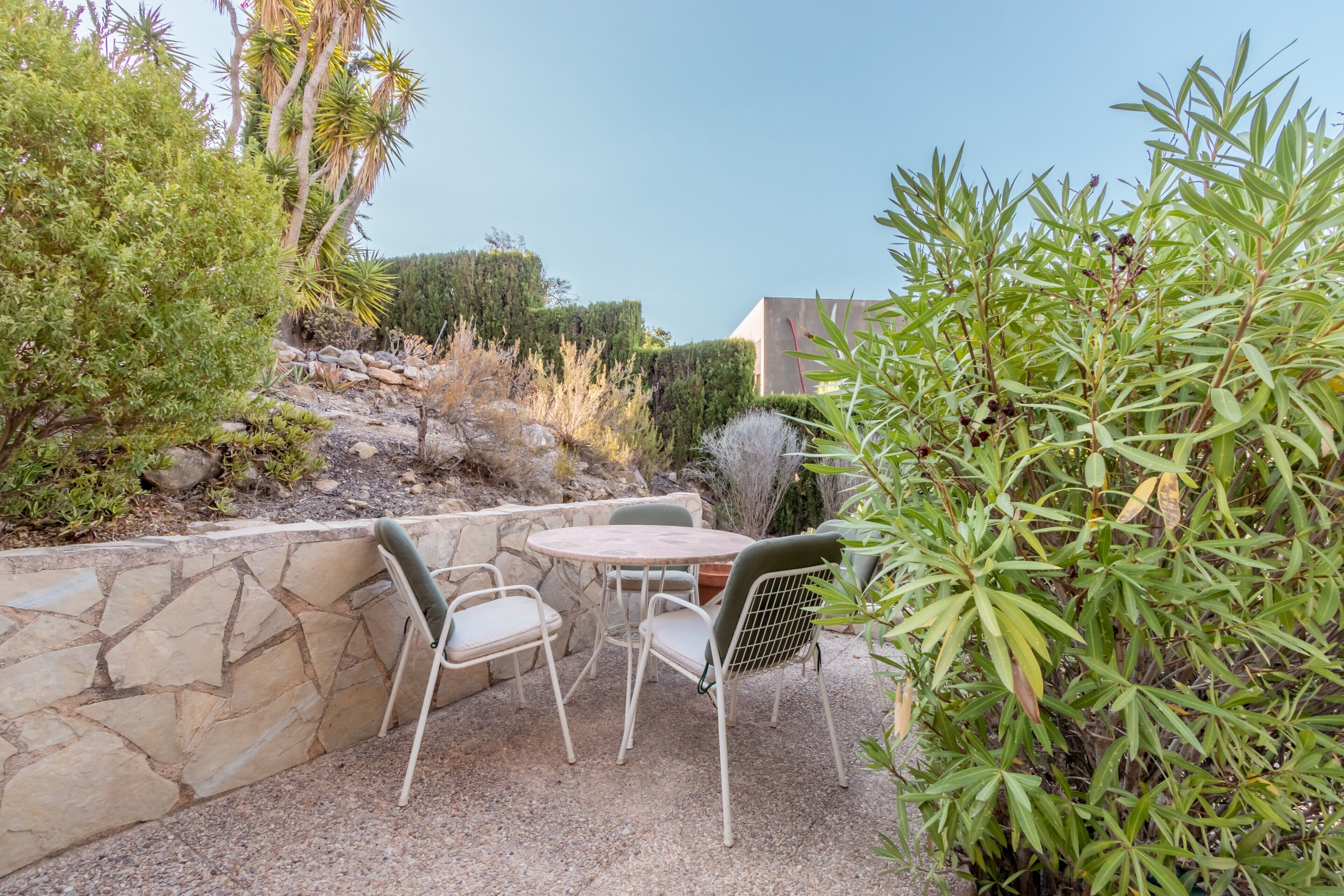 Un patio con pared de piedra, dos sillas, una mesa y abundante vegetación.