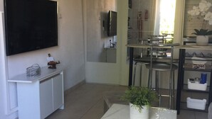 Ein modernes Wohnzimmer mit Fernseher, einem kleinen weißen Tisch mit Pflanze und einem durchgegangene Tür erkennbaren Küchenbereich.
