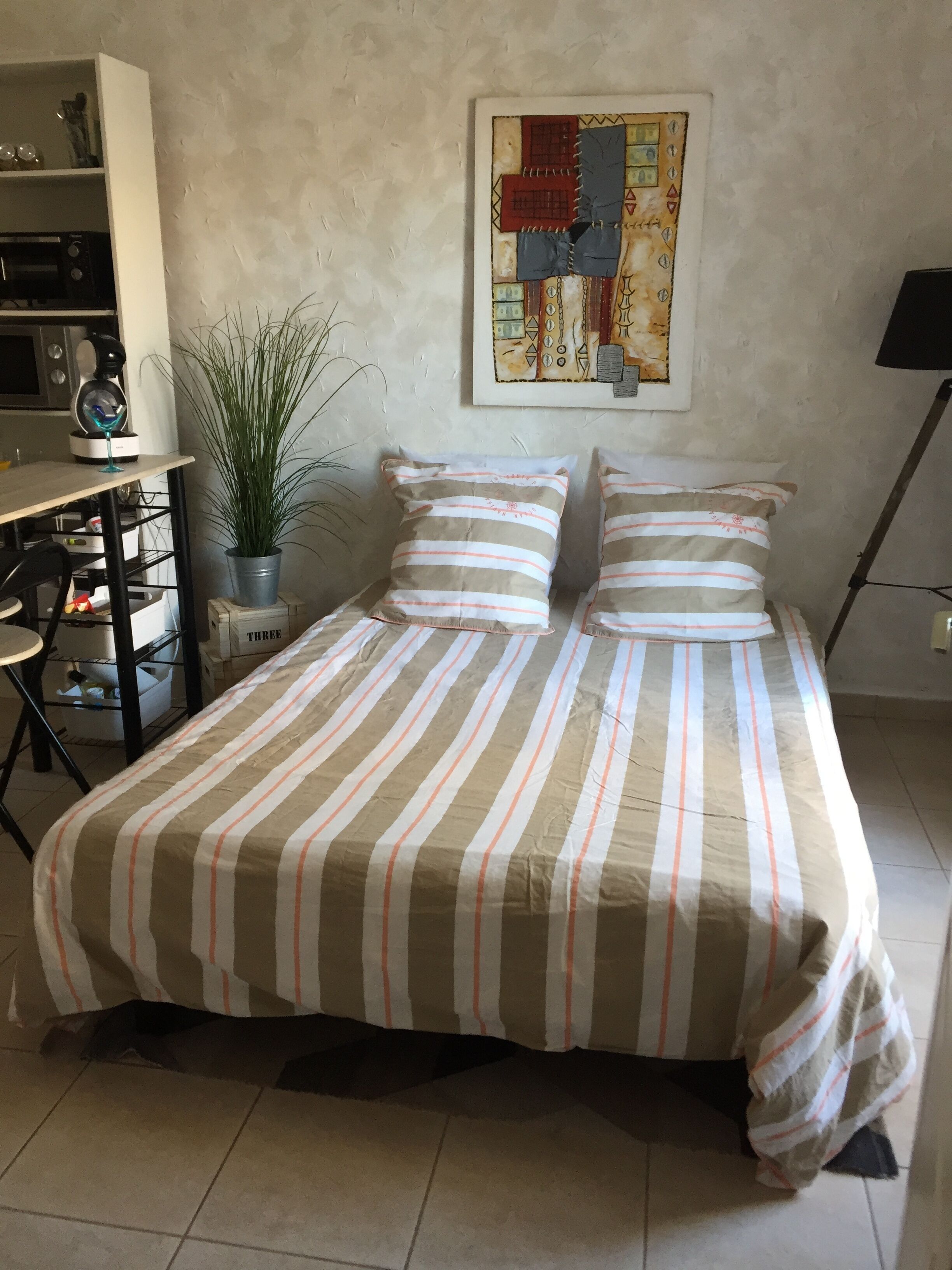 A bedroom with a bed, a desk, a chair, a potted plant, and a painting on the wall.