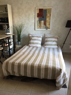 A bedroom with a bed, a desk, a chair, a potted plant, and a painting on the wall.