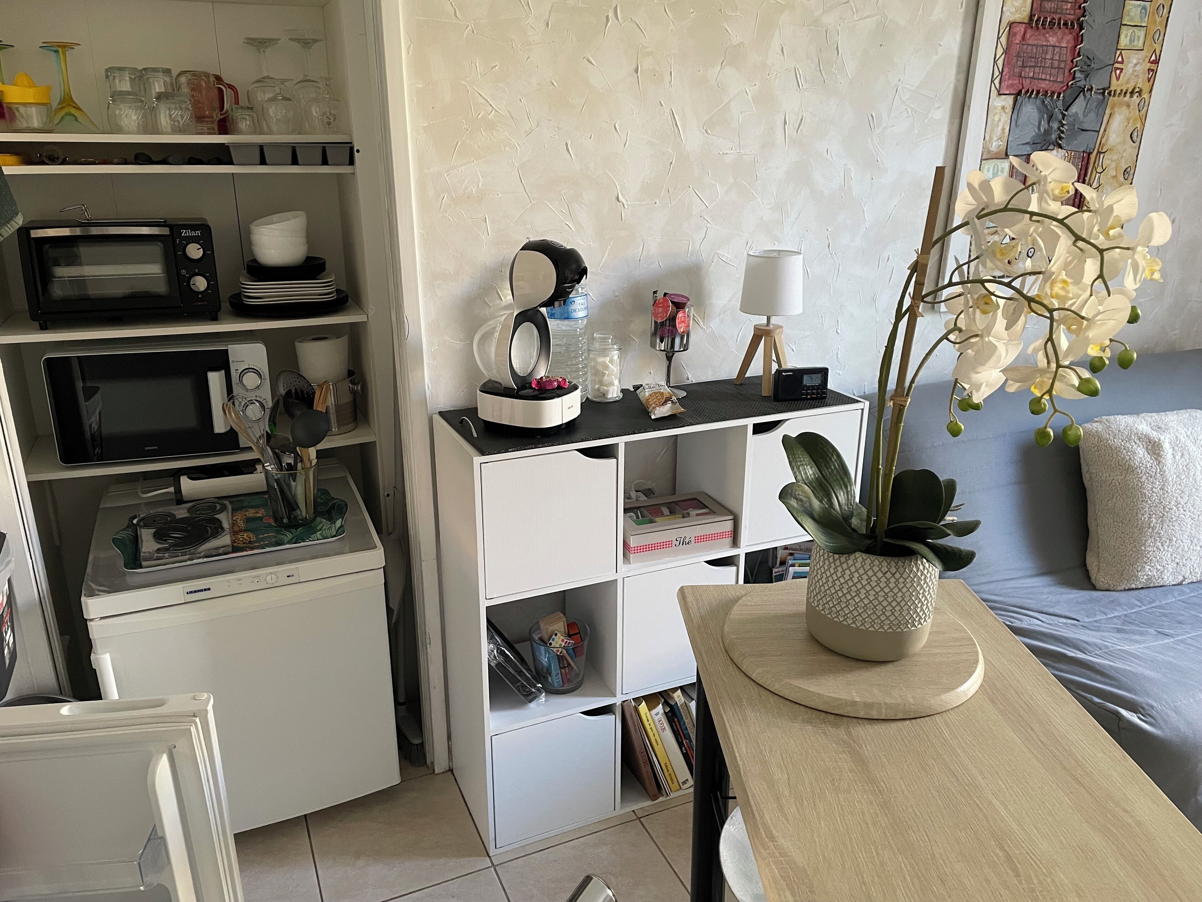 A compact kitchen with a microwave, toaster, and kettle. There's a small dining area with a table and chairs, and a bookshelf filled with books and decorative items.
