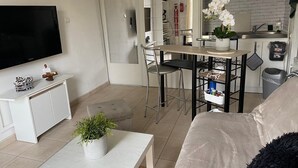Ein modernes Wohnzimmer mit Flachbildfernseher, einem weißen Sofa, einem Couchtisch mit Pflanze und einer Küchenzeile mit Spüle und Orchidee im Topf.