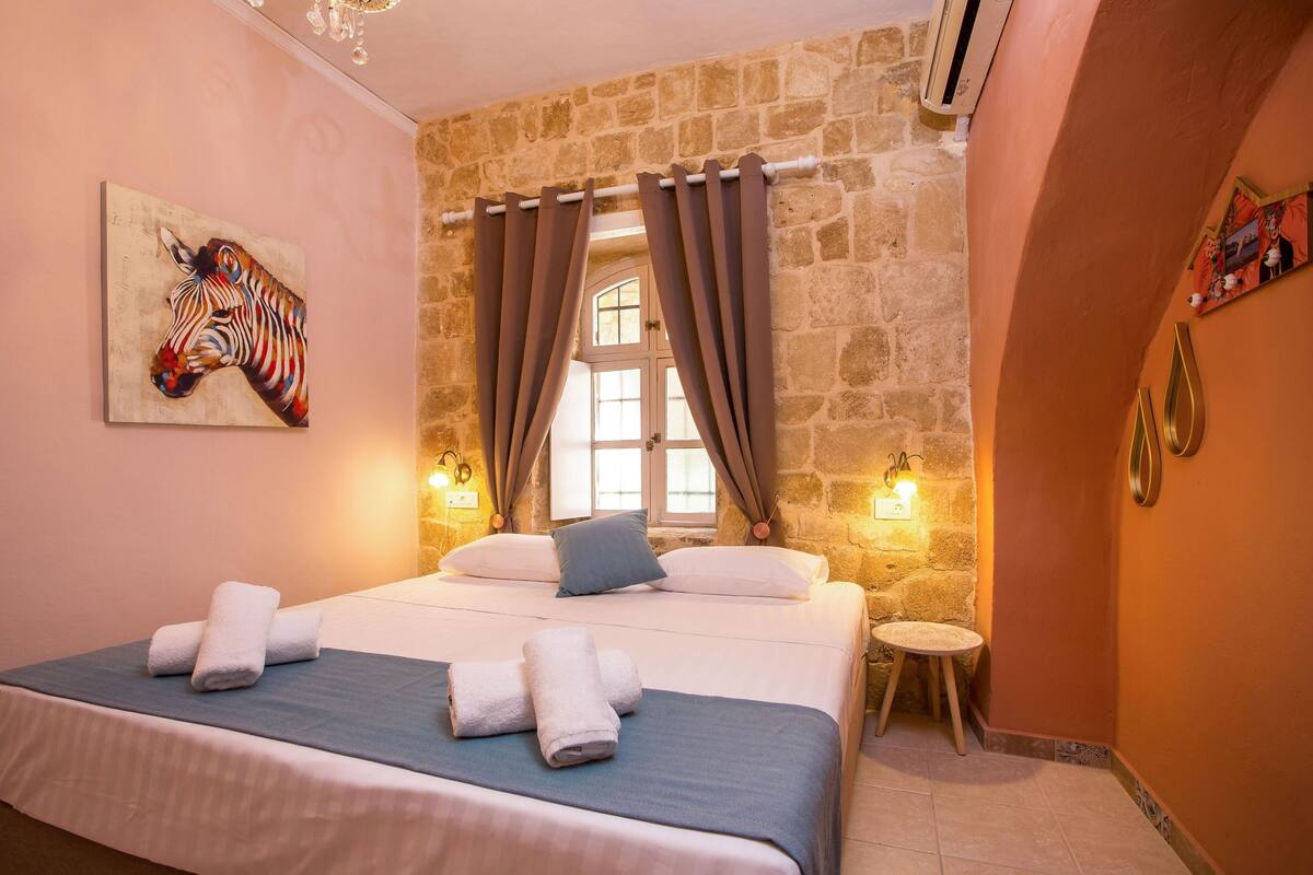 A bedroom with a stone wall, a bed with towels, a window with curtains, and wall-mounted lights.