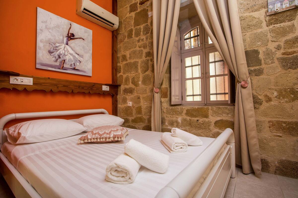A bed with white linens, a painting of a dancer, stone walls, and a window with curtains.