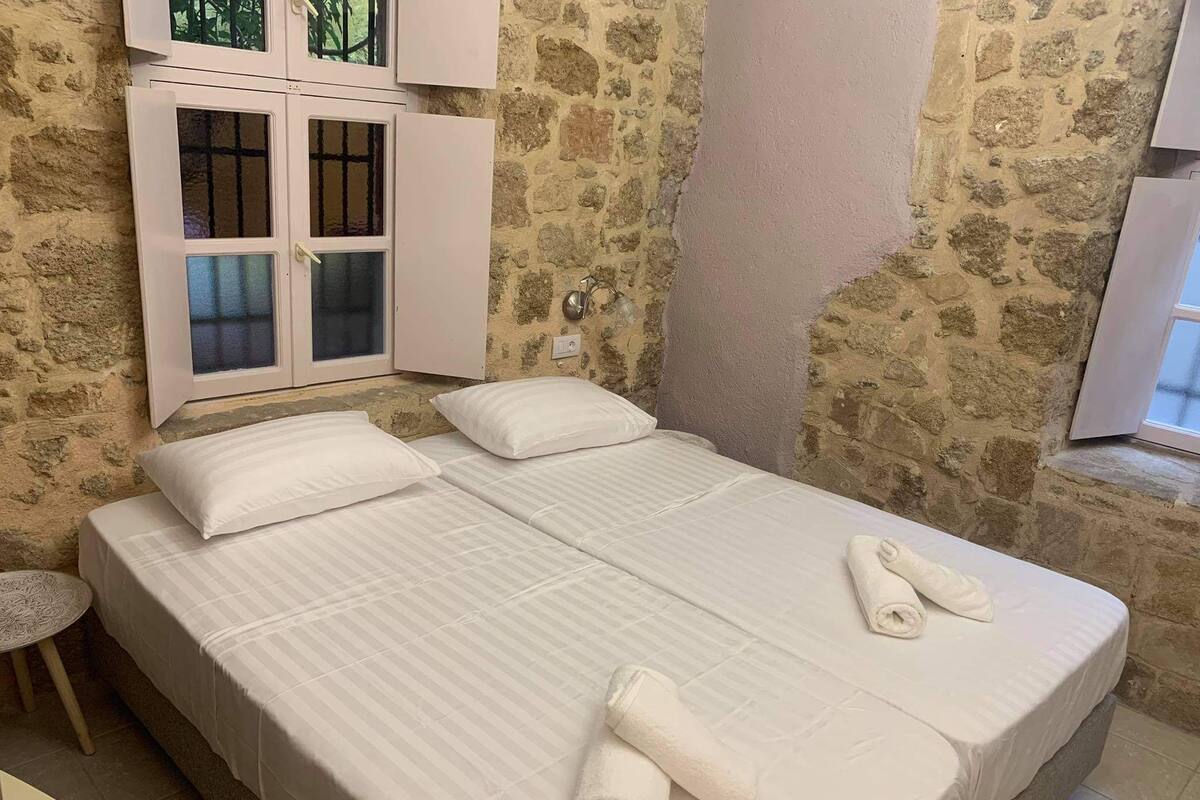 A double bed with white linens and two rolled towels, a stone wall, and a window with a view.