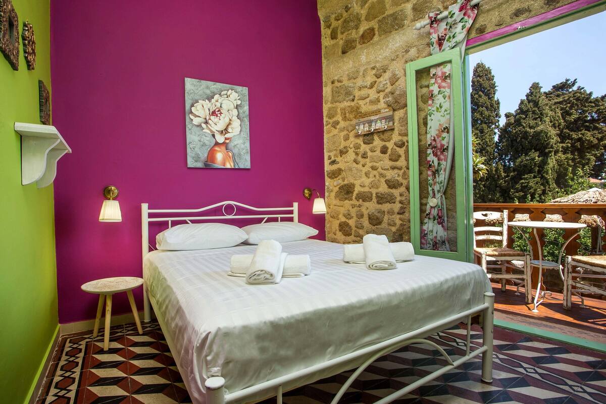 A bedroom with a bed, pillows, a nightstand, a small table, and a painting on the wall.