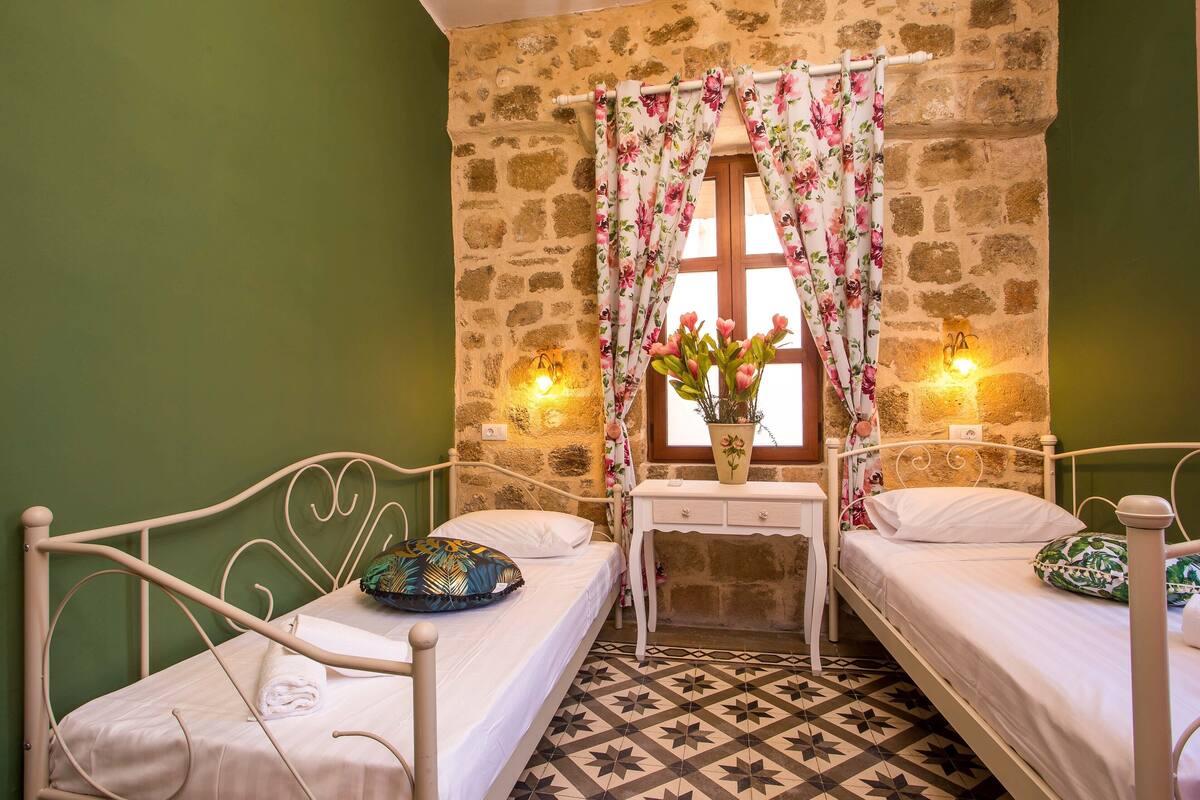 A room with two beds, a stone wall, a window with floral curtains, a small table with a vase, and two wall-mounted lights.