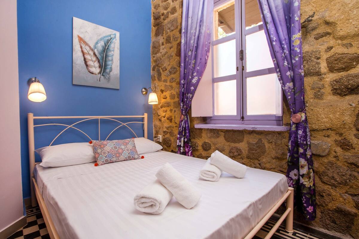 A bed with white linens, a patterned pillow, and rolled towels. There is a window with purple curtains and a stone wall.