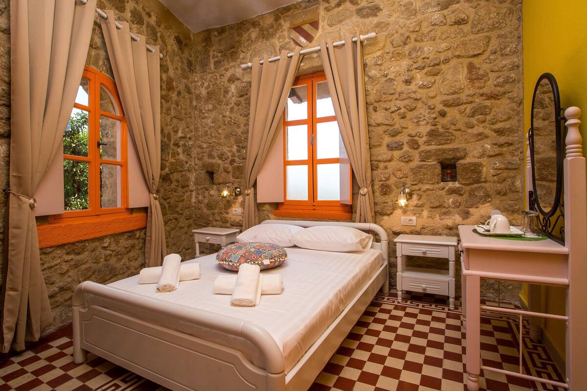 A bedroom with a stone wall, a checkered floor, a bed with white bedding, a bedside table, and a small desk with a mirror.