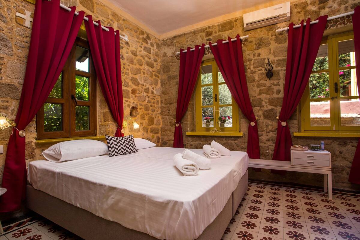 A bedroom with a stone wall, a bed with white linens, red curtains, a bench, and a small bedside table with a bottle and a book.