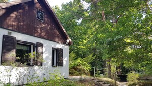 A rustic wooden house with white walls and dark shutters, surrounded by lush greenery and a stone pathway.