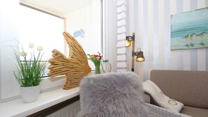 Un accogliente soggiorno con termosifone bianco, una poltrona grigia in pelo, un divano beige, un oggetto decorativo in legno, una pianta in vaso e un vaso con fiori.