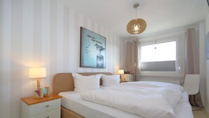 Un letto rifatto con lenzuola bianche, un comodino con una lampada, un quadro incorniciato sulla parete e una finestra con tende.