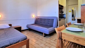 Een compacte kamer met een bed, een slaapbank, een eettafel met stoelen en een kitchenette.