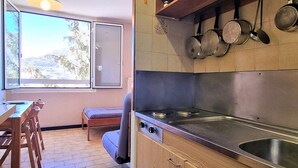 Een keuken met een kookplaat, gootsteen en hangende pannen. Een raam biedt uitzicht op bomen en heuvels.