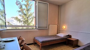 Een kleine, goed verlichte kamer met een enkel bed, een tafel met stoelen en een raam met uitzicht op bomen en bergen.