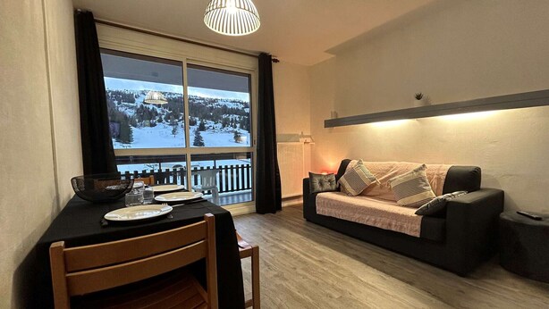 A cozy living room with a dining area, a sofa, and a view of a snowy landscape through large windows. - Comfortable studio for 4 with terrace, parking, wifi and pets allowed (Le Dévoluy)