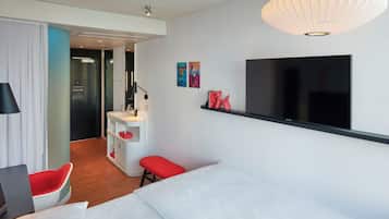 Una habitación de hotel moderna con una cama, un escritorio con silla, un televisor y un banco rojo.