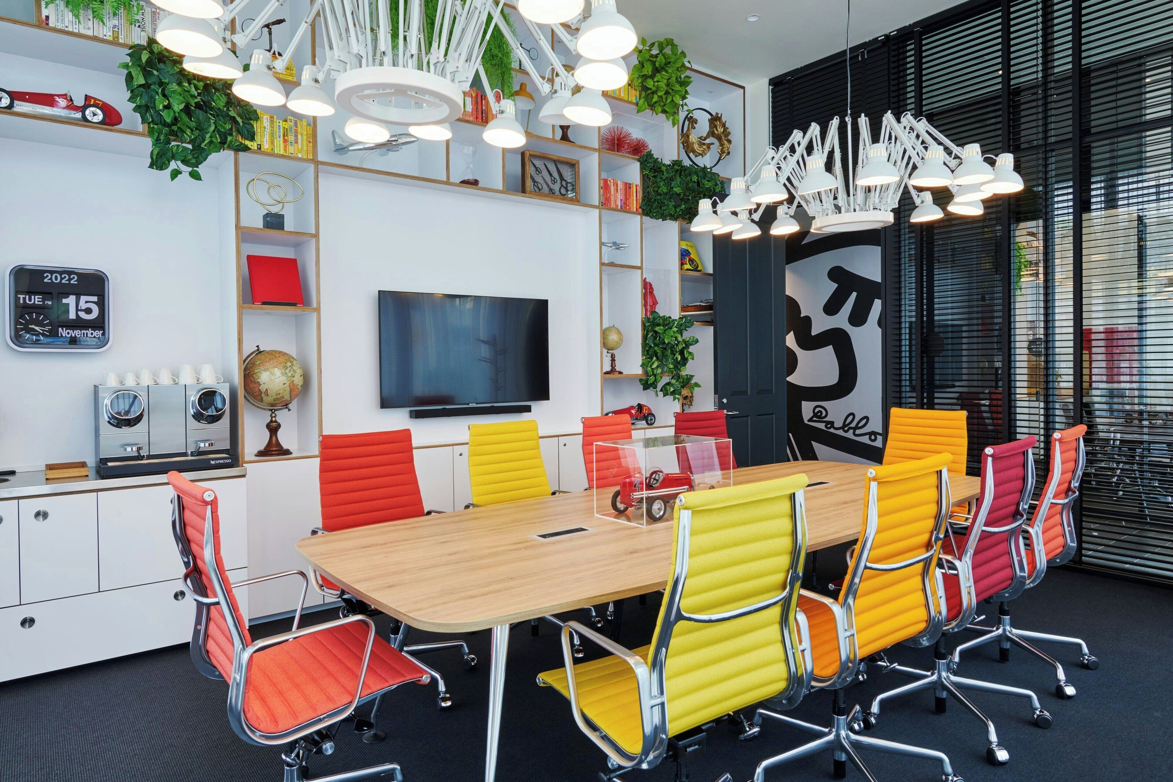 Una sala de reuniones moderna con una mesa de madera, sillas coloridas, un televisor grande y varios objetos decorativos en las paredes.
