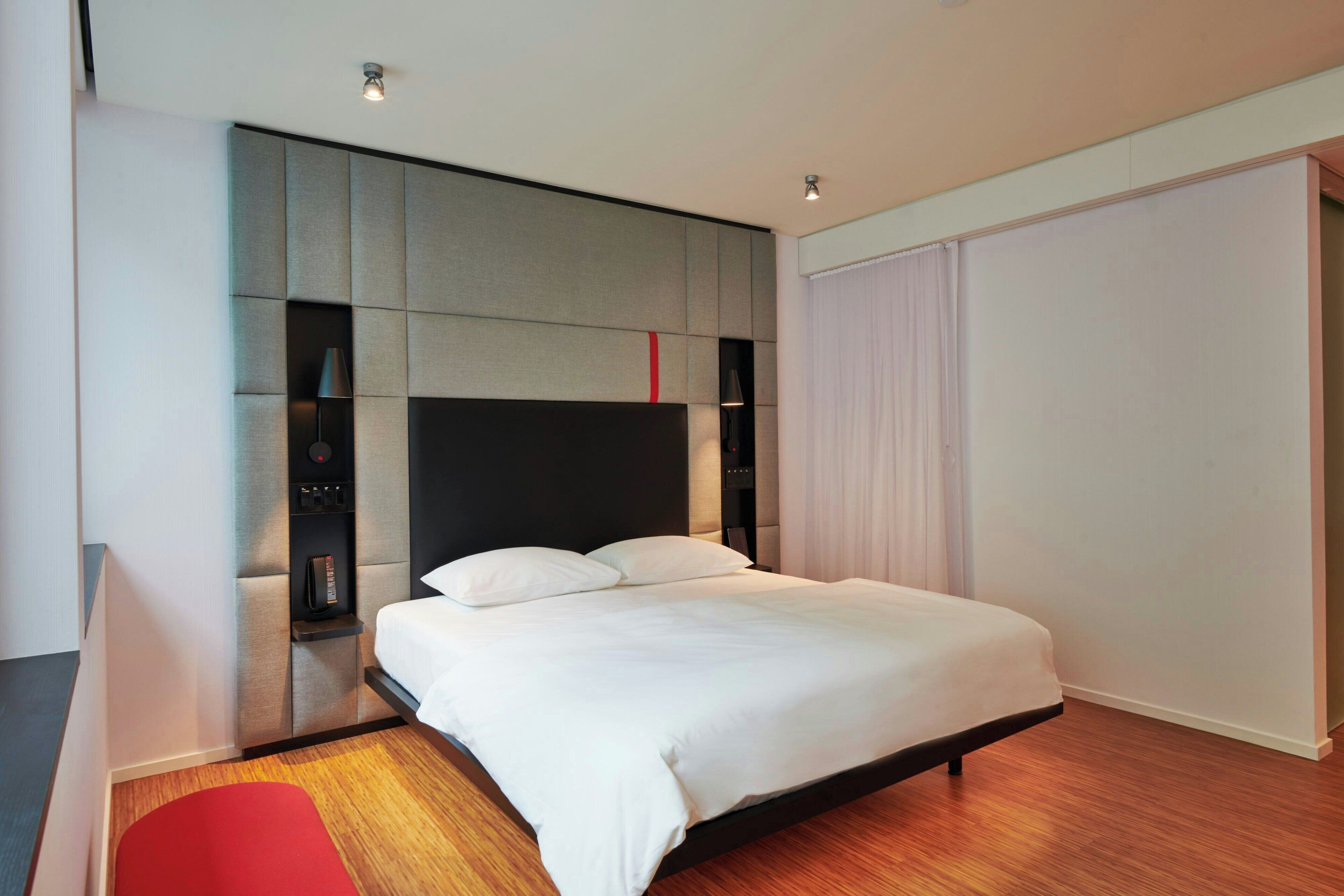 Una habitación de hotel moderna con una cama grande, almacenamiento integrado y una butaca roja.