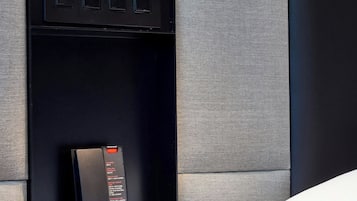 Hotellihuone, jossa on puhelin sängyn vieressä, valaistuksen säätöpaneeli ja pieni hylly.