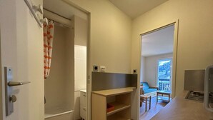 A compact room with a shower, a small desk, a chair, and a view of a balcony.