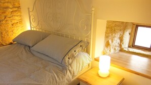 A bedroom with a bed, a nightstand, a lamp, a stone wall, and a window.