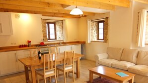A cozy living room with a wooden dining table, chairs, and a sofa. A kitchen with stone walls and wooden beams is visible in the background.