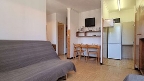 A compact living space with a sofa, a dining table with chairs, a kitchen area with a refrigerator and a microwave, and a TV mounted on the wall.