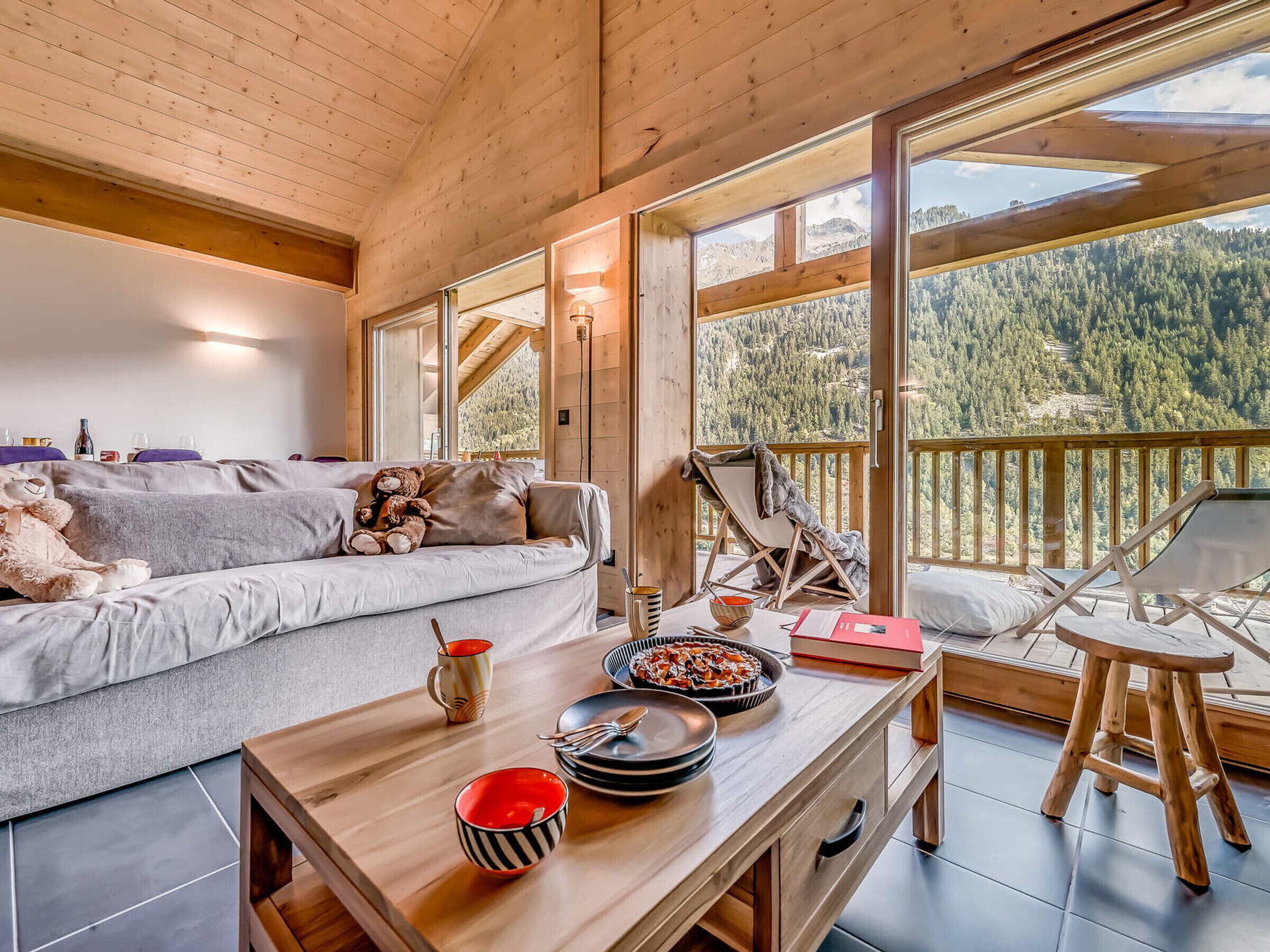 Interior acogedor de una cabaña con techo de madera, un sofá con cojines, una mesa de comedor con platos y cuencos, y vista a una cadena montañosa boscosa a través de grandes ventanales.