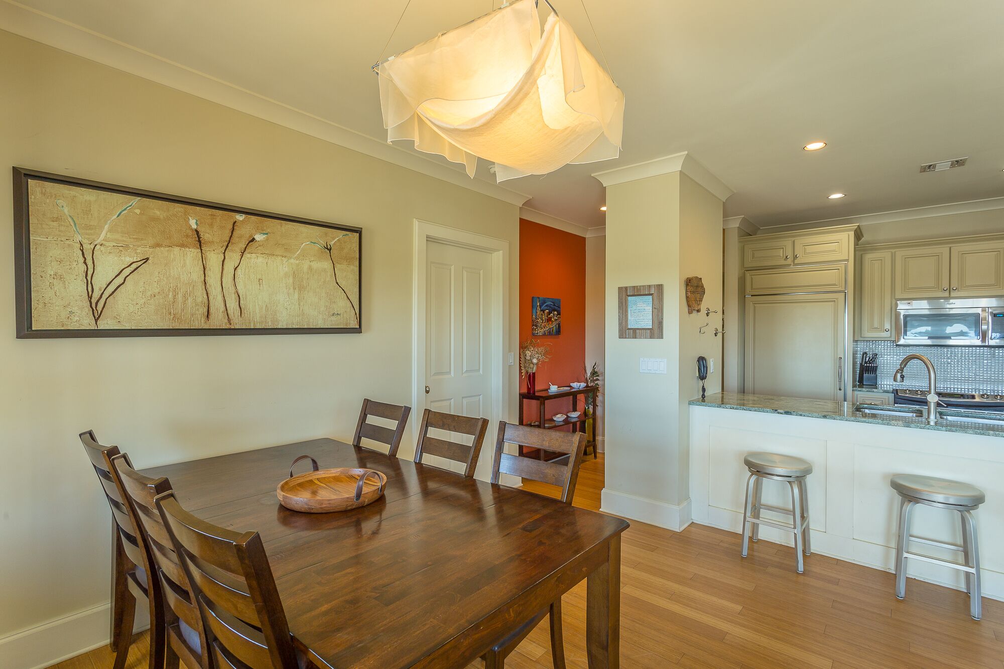 A dining area with a wooden table and chairs, a hanging light fixture, and a kitchen with stainless steel appliances.