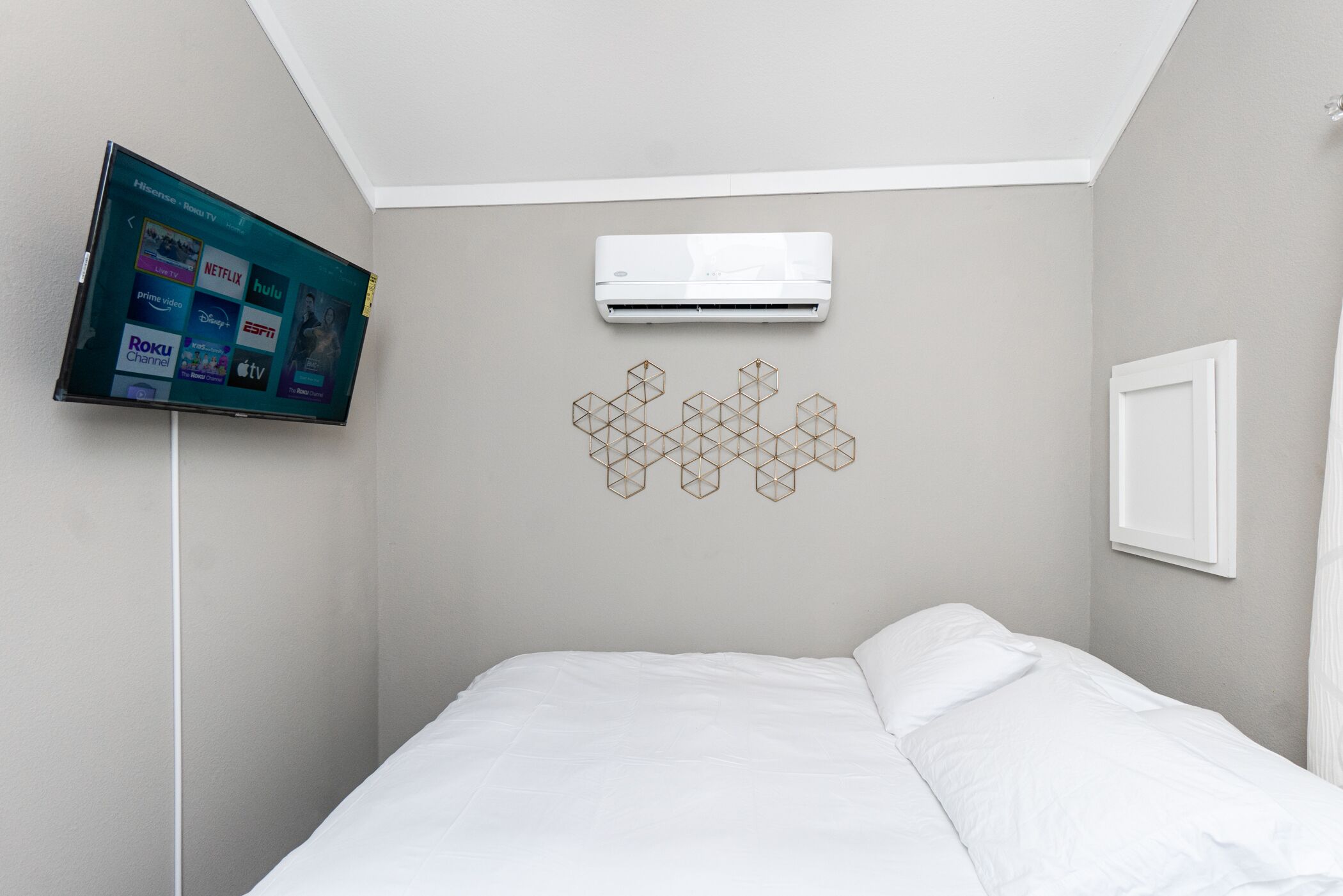 A bedroom with a bed, a mounted TV displaying streaming services, an air conditioner, and a geometric wall decoration.