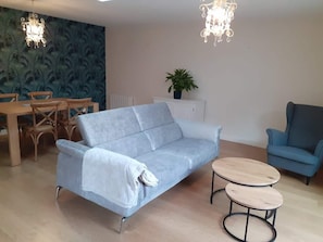 Una sala de estar con un sofá azul claro, un sillón azul, una mesa de comedor de madera con sillas y dos mesas redondas de madera.
