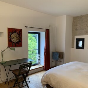 Une chambre à coucher avec un lit, un bureau avec une chaise, une fenêtre donnant sur un paysage verdoyant et un tableau accroché au mur.