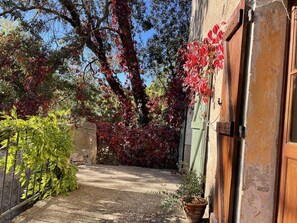 Une ruelle étroite avec un chemin de gravier, bordée d’arbres aux feuilles rouges, d’un mur de pierre et d’une porte en bois.
