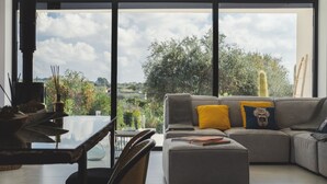 Ein modernes Wohnzimmer mit großem Fenster, einem grauen Sofa mit gelb-blauen Kissen, einem Glastisch und Blick auf Grünflächen und Bäume.