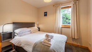 Een keurig opgemaakt bed met witte lakens, een nachtkastje met een lamp en een raam met gordijnen.