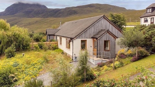 Een rustiek huis met een schuin dak, een klein terras en een tuin met diverse planten.