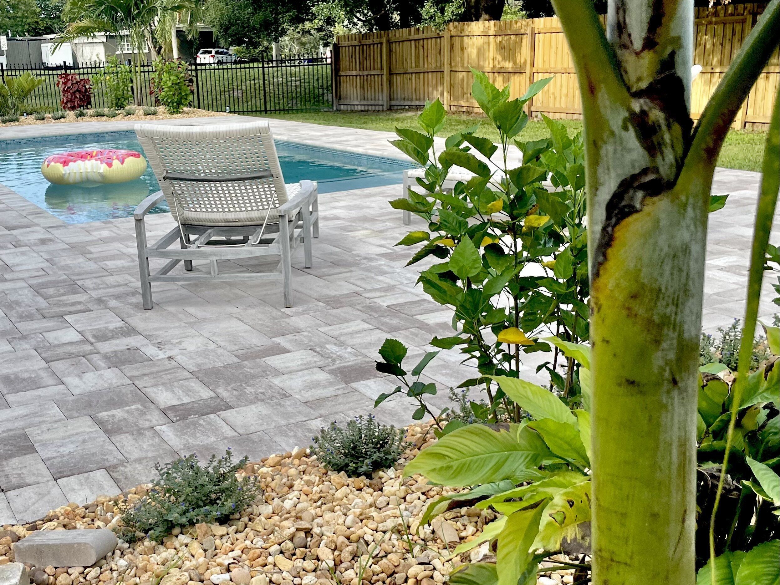 Un patio trasero con piscina, una silla de descanso y un jardín con plantas y piedras.