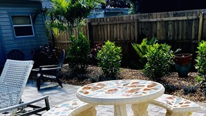 Una mesa de patio con mosaicos y cuatro sillas, rodeada de baldosas de piedra y plantas en macetas.