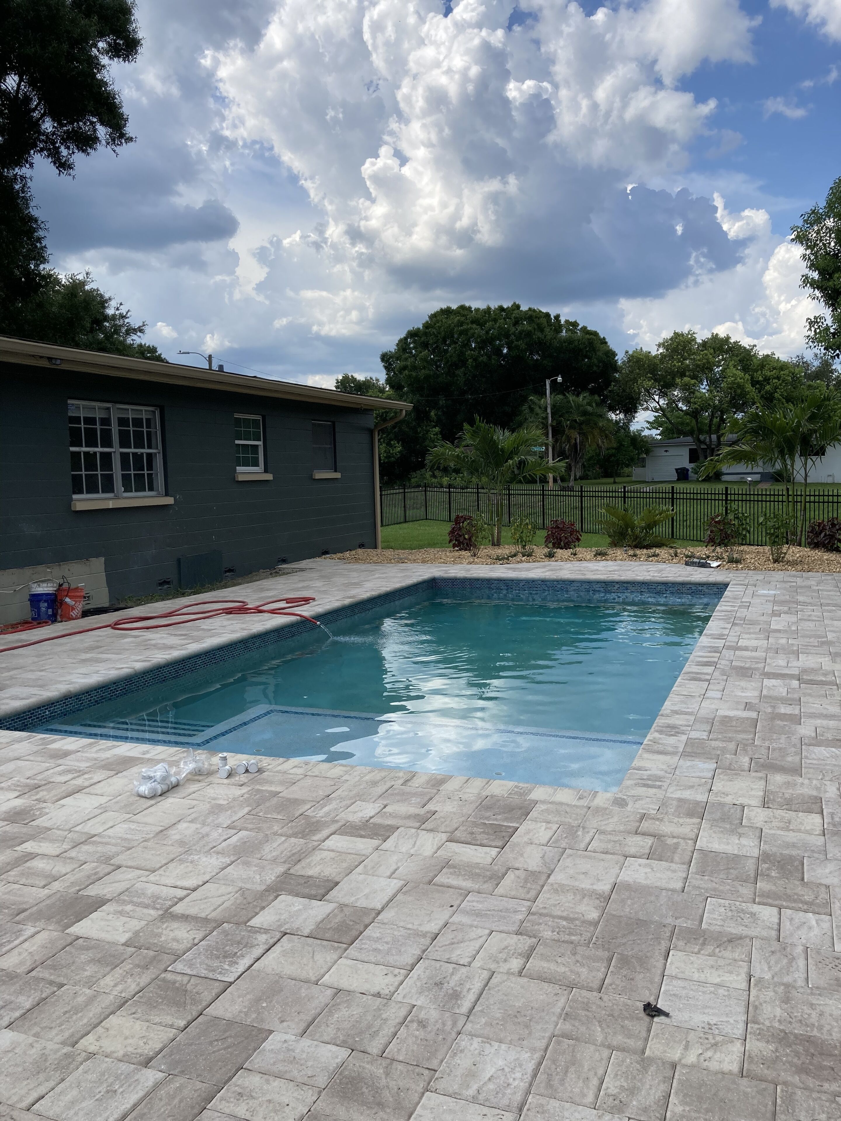 A swimming pool with a tiled deck, a small house, and a fenced yard with trees.