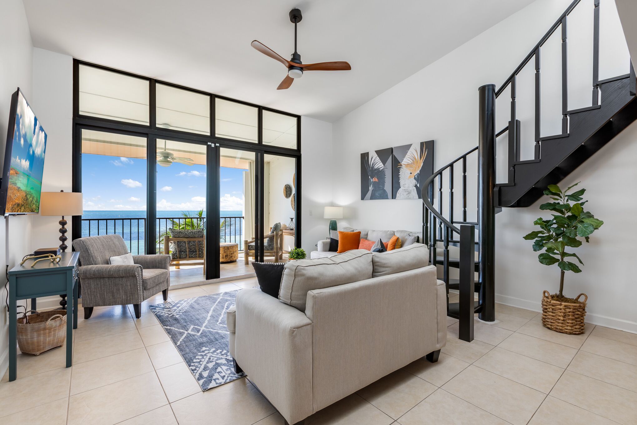 A spacious living area with a large window, a ceiling fan, a staircase, and a view of the ocean.