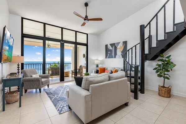 A spacious living area with a large window, a ceiling fan, a staircase, and a view of the ocean.