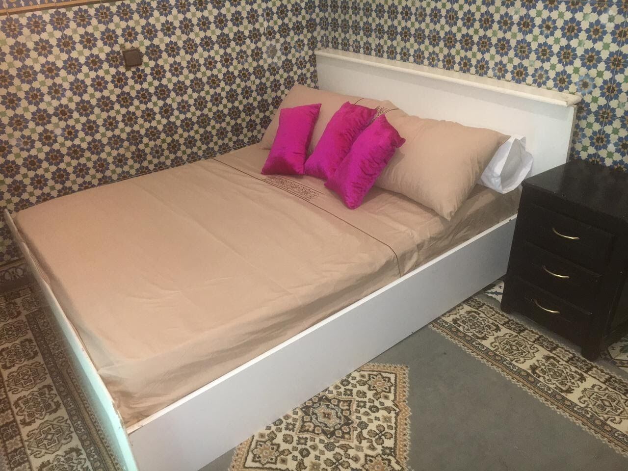 Una cama con colchón color beige y almohadas rosas, cabecera blanca y mesita de noche oscura con cajones.