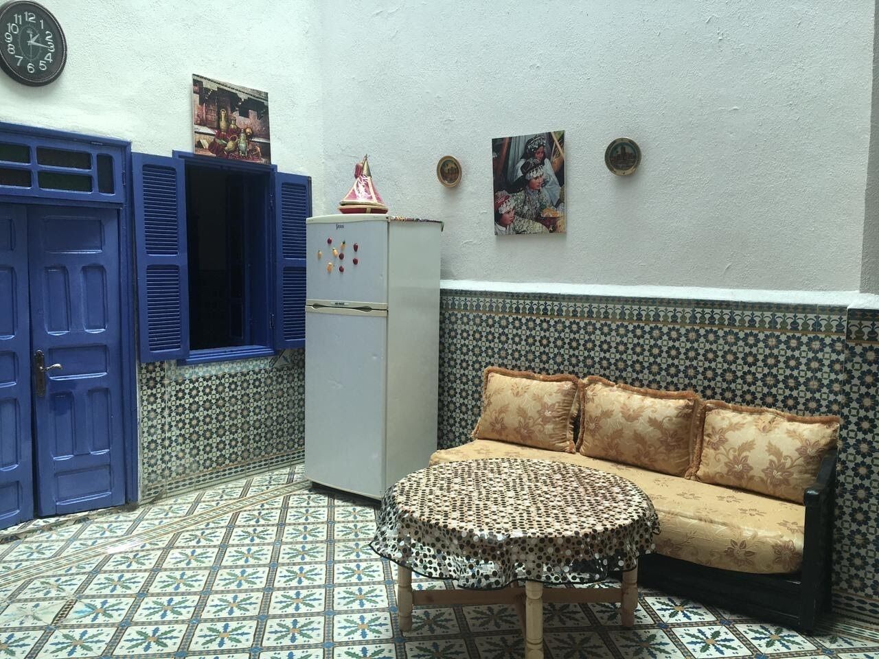 A room with a patterned floor, a sofa with floral cushions, a small table, and a refrigerator.