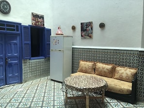 Ein Zimmer mit einem gemusterten Fliesenboden, einer Couch mit floralen Kissen, einem kleinen runden Tisch, einem Kühlschrank und blauen Türen.