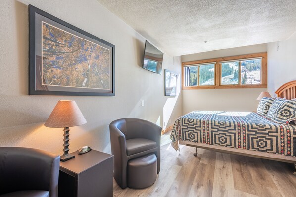 A bedroom with a bed, a chair, a desk, a lamp, and a large framed picture on the wall.
